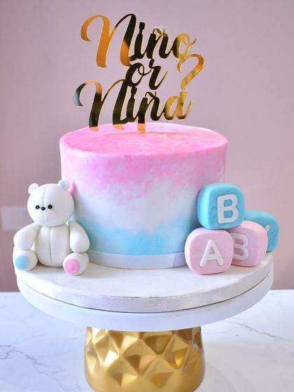 A lovely gender reveal cake with a "Niño or Niña?" theme. The cake has a pink and blue ombre effect and is decorated with a sweet teddy bear and fondant baby blocks.