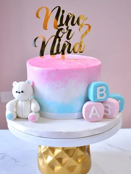 A lovely gender reveal cake with a "Niño or Niña?" theme. The cake has a pink and blue ombre effect and is decorated with a sweet teddy bear and fondant baby blocks.