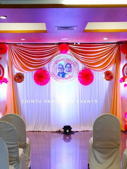 A grand stage decoration for a 50th wedding anniversary. The setup uses orange and white drapes, paper fans, and a personalized photo of the couple.