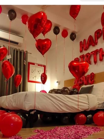 A dramatic and romantic birthday room decoration. The room is filled with red and black balloons, including heart-shaped foils, with 'HAPPY BIRTHDAY' letters on the wall and a floor covered in rose petals.