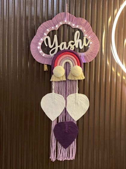 A beautiful nameplate for 'Yashi' in a lovely lilac and purple color scheme. The design includes a rainbow, pom-pom clouds, and hanging macrame leaves, all illuminated by soft fairy lights.