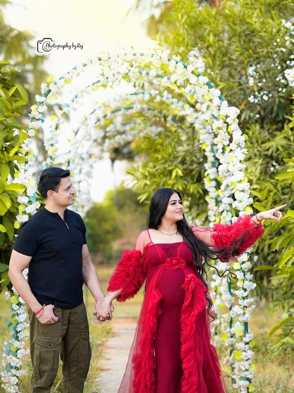 A candid moment of a couple walking through a floral archway. The mother-to-be is wearing a beautiful red ruffled gown.