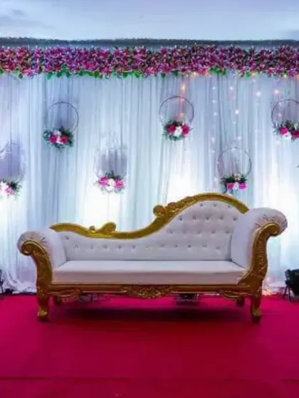 A simple and elegant reception stage with a white backdrop. The decor features hanging floral hoops and a classic white diwan, creating a minimalist yet beautiful setting.
