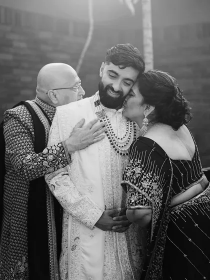 A touching black and white photo of a groom receiving a kiss from his mother and a hug from his father. This intimate family moment is full of love and pride.