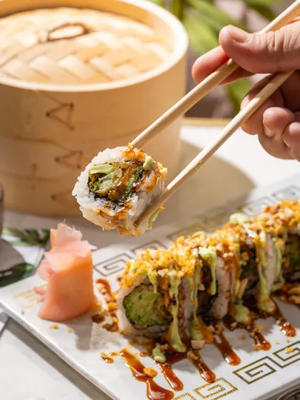A close-up of a crunchy avocado sushi roll being lifted by chopsticks. The focus is on the texture of the tempura flakes and creamy sauce.