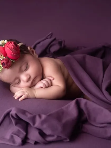 The simplicity of a sleeping newborn on a rich purple backdrop. The floral headband adds a delicate pop of color, highlighting the baby's beautiful features.