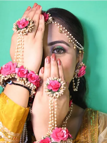 An artistic close-up of a model wearing a complete floral jewellery set. This shot highlights the hath phool (hand harness), rings, and maang tikka made of beautiful pink roses and pearls.
