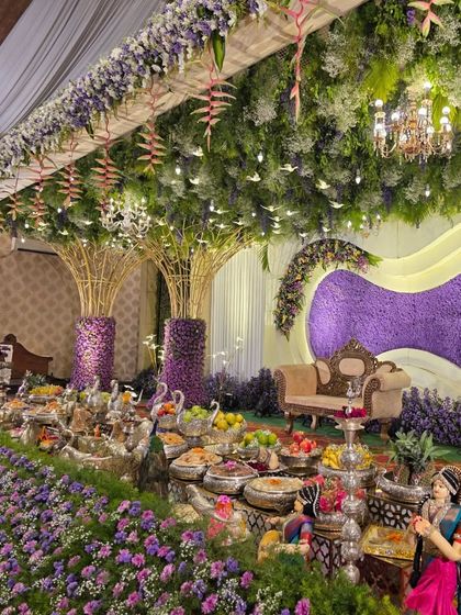 An angled view of the purple floral stage, highlighting the hanging greenery, chandeliers, and the detailed arrangement of silver props and offerings in the foreground.
