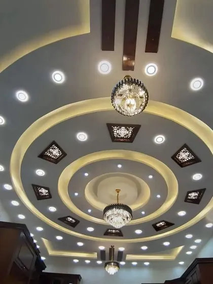 A magnificent circular ceiling design perfect for a large hall or living room. It features multiple layers, two chandeliers, and numerous spotlights, creating a truly grand and brightly lit space.