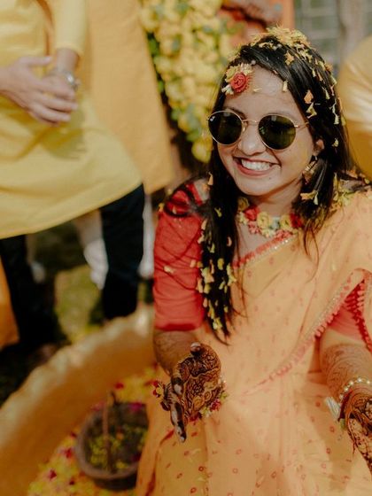 The bride, wearing sunglasses and a huge smile, is ready for the Haldi fun. This shot captures her cool, happy vibe amidst the flower petals and yellow decor.