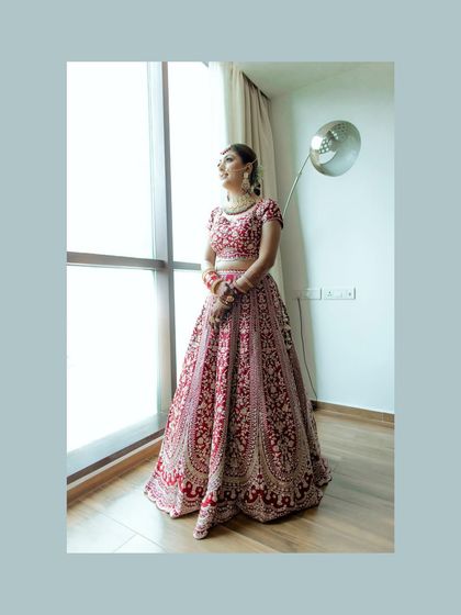A full-length bridal portrait by the window. The natural light beautifully illuminates her red lehenga and her hopeful expression as she looks outside.