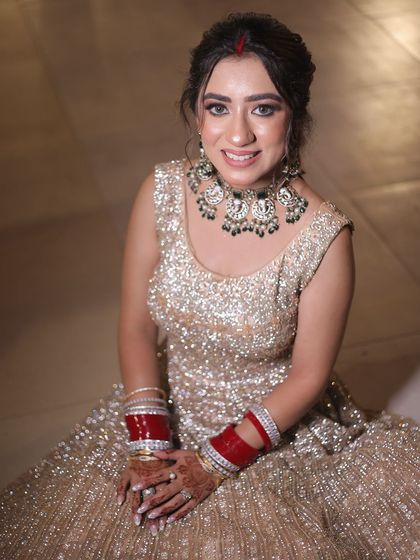 A portrait of a bride seated, looking directly at the camera, showcasing her sparkling gown and traditional red bangles.