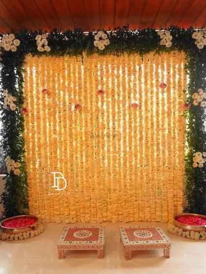 This is a classic and grand backdrop for a Haldi or mehndi ceremony, made entirely of marigold strings. The setup is framed with green foliage and includes traditional low seating stools, perfect for rituals and photos.