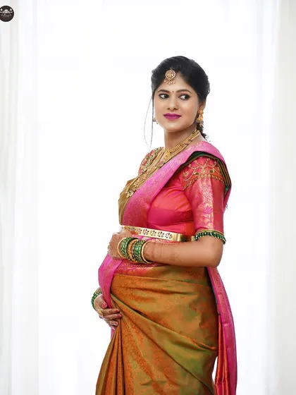 This studio portrait captures the expectant mother in a classic pose. The simple white backdrop ensures that the vibrant colors of her saree and her joyful expression are the main focus.
