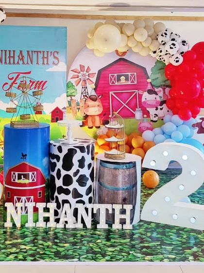 A charming farm-themed second birthday party. This setup includes a red barn backdrop, cow-print plinths, a white picket fence, and cutouts of farm animals like cows and sheep, bringing a touch of the countryside to the celebration.