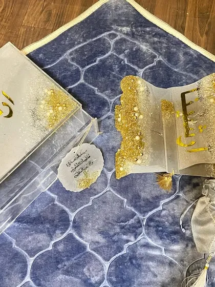 A personalized gift set featuring a dome-shaped prayer mat in a beautiful blue pattern. The set is completed with a custom resin Quran box and holder, a crystal tasbeeh, a bookmark, and a car hanging, all in a coordinated white and gold glitter design.