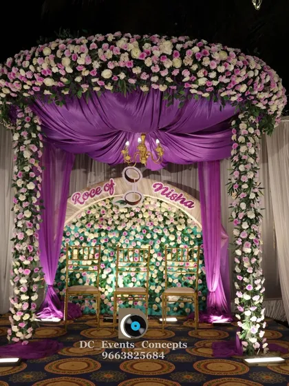 A grand and regal Roce ceremony setup featuring a canopy of purple fabric and flowers. The flower wall backdrop and elegant chairs create a luxurious and traditional atmosphere for the ceremony.