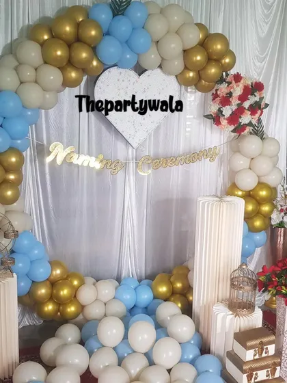 A simple and elegant naming ceremony decoration with a white, blue, and gold balloon arch. The heart-shaped cutout and "Naming Ceremony" banner add a sweet touch.
