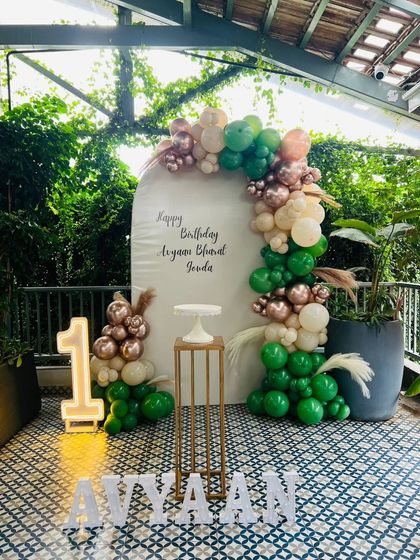 A chic woodland theme for a first birthday, set up on a patio. This design uses an arched white backdrop with custom calligraphy, a balloon garland in green, beige, and rose gold, and a light-up number '1'.
