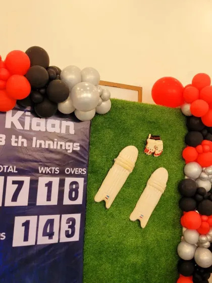 A close-up of the custom scoreboard and cricket pad props on the artificial grass backdrop.