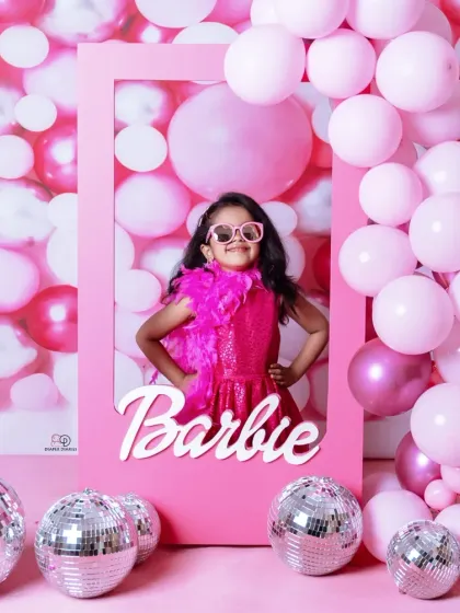 A vibrant 'Barbie' themed photoshoot for a stylish toddler. She poses confidently in a pink frame against a wall of pink balloons and disco balls, complete with a feather boa and sunglasses.