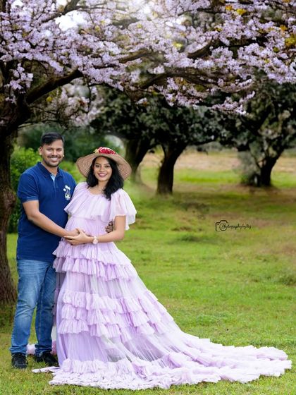 A lovely couple's portrait under blooming trees. The mother-to-be is wearing a lavender ruffled gown and a hat.