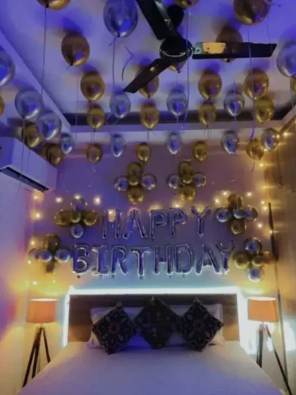 A simple birthday room decoration with silver and gold balloons on the ceiling and "HAPPY BIRTHDAY" foil letters on the wall.