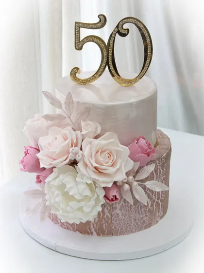 Cheers to 50 years! This anniversary cake combines a textured rose gold base with a smooth pearlescent top tier, adorned with a cascade of handmade sugar flowers in soft pinks and whites.