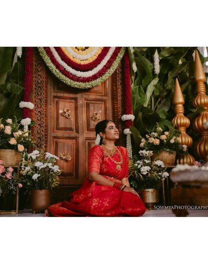 A bride in a vibrant red saree seated before a beautifully decorated floral backdrop, looking serene and elegant.
