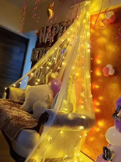 A cozy corner decorated for a birthday at home, with a chair draped in a fairy light canopy and surrounded by balloons.