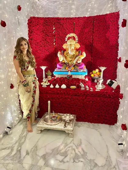 The client looking radiant in her traditional attire, posing next to the stunning red rose Ganpati mandap. This shows how the decor creates a perfect photo opportunity.