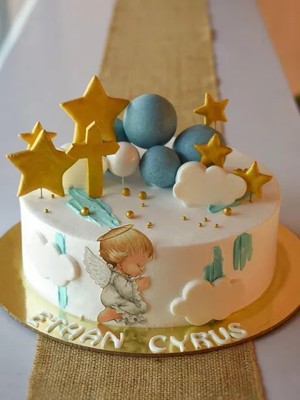 A sweet and serene whipped cream cake for baby Ethan's baptism. The design features a praying angel, fondant clouds, and gold stars in a gentle color palette of blue, white, and gold.