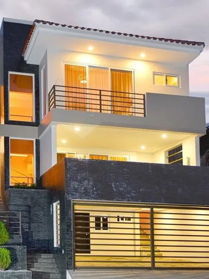 This modern house is designed for a sloped plot, with a garage at the lower level and living spaces above. The exterior uses a dark stone base and white upper floors, with strategic lighting to highlight its multi-level architecture at night.