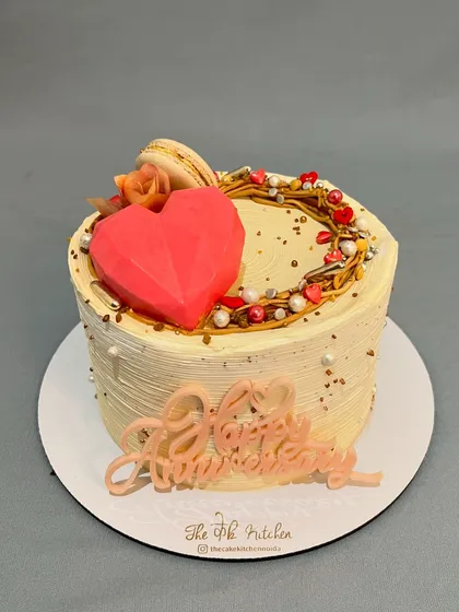 This lovely anniversary cake is topped with a geometric red heart and a delicate wreath of buttercream and sprinkles. A single macaron and a custom 'Happy Anniversary' sign complete this sweet and romantic design.