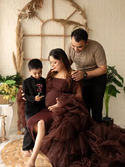 A heartwarming family moment in the boho setup. The older brother points excitedly at his mother's baby bump as his father looks on with a smile.