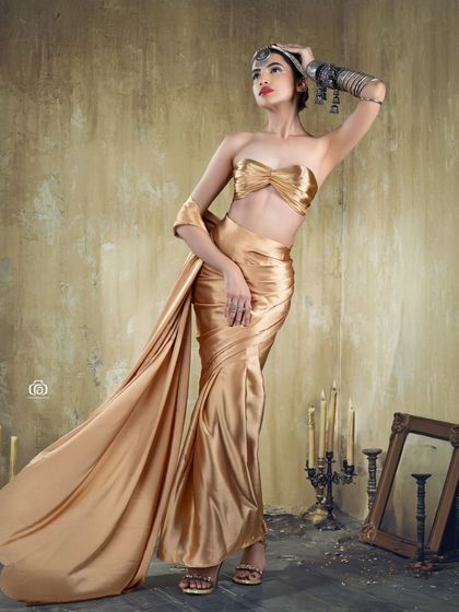 A full-length studio shot of a conceptual golden goddess look. The flowing fabric, antique-style props, and warm lighting combine to create a dramatic and artistic fashion portrait.