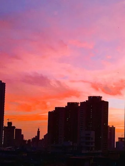 A vibrant, fiery sunset with shades of pink, purple, and orange creates a dramatic backdrop for the silhouetted Mumbai skyline.