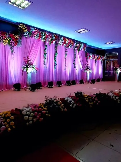 This wide-angle view showcases a complete stage setup with a multi-layered floral border and cascading greenery. The purple and pink lighting scheme adds a vibrant and celebratory feel to the entire hall.