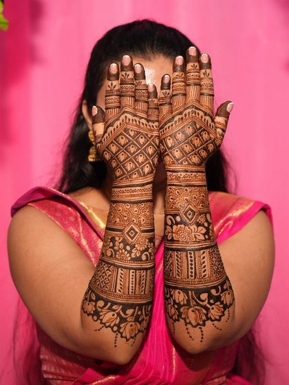 A classic bridal pose against a pink backdrop, with the bride's hands hiding her face to showcase the henna.