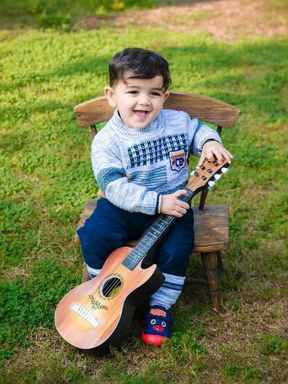 A big, happy smile from a toddler boy holding a guitar during his outdoor photoshoot. His joy is evident.