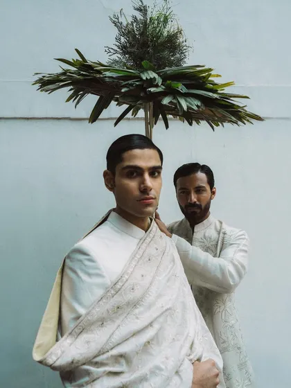 A conceptual shot for Sanchit Sharma, featuring one model adjusting the other's garment under a leafy parasol. The image tells a story of preparation and quiet intimacy.