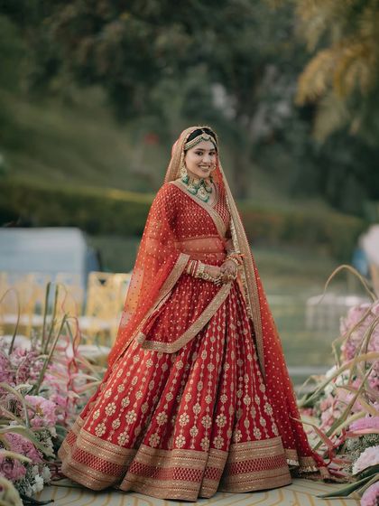 A classic bridal portrait of Rhythm at her Udaipur wedding. The vibrant red of her Sabyasachi lehenga stands out beautifully against the lush greenery and floral decor of the Taj Aravali.