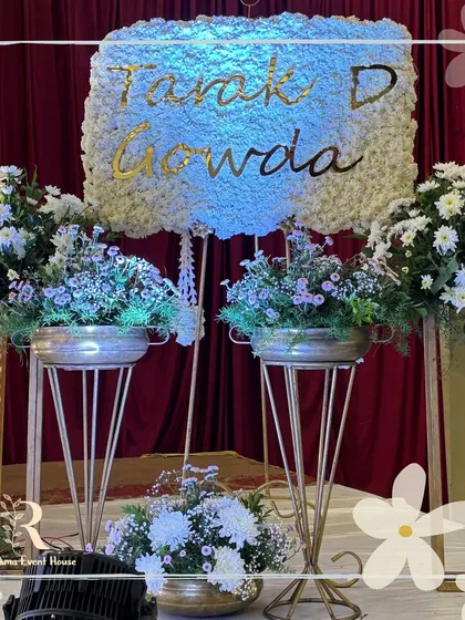 A creative name reveal setup for "Tarak D Gowda." The name is elegantly scripted in gold on a dense bed of white flowers, surrounded by fresh floral arrangements in brass pots, making the reveal a beautiful moment.