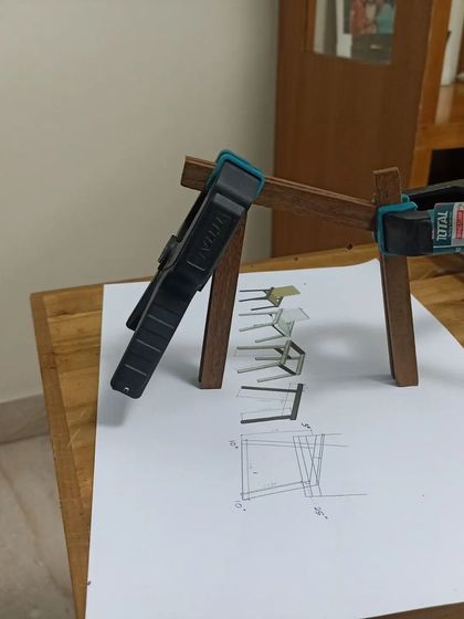 Using a clamp to hold the miniature chair frame together while the glue sets. The design sketch in the background shows the reference for the model.