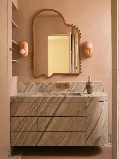 We call this bathroom a 'chic desert dream'. It features blush-toned micro-concrete walls and a monolithic vanity carved from veined Corteccia marble. The stone appears to trickle down onto the floor, creating a fluted skirting detail that is both organic and luxurious.