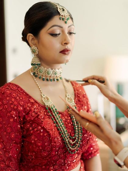 A behind-the-scenes moment, applying the finishing touches. The combination of the red outfit and green jewelry is a classic and beautiful choice.