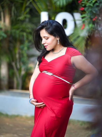 A classic portrait of a mom-to-be in a striking red dress, looking down at her baby bump in a garden setting.