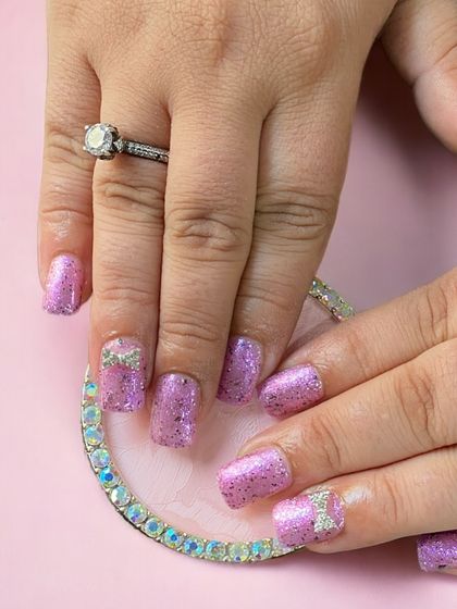 A fun and sparkly purple glitter set on shorter square nails, with cute 3D rhinestone bows.
