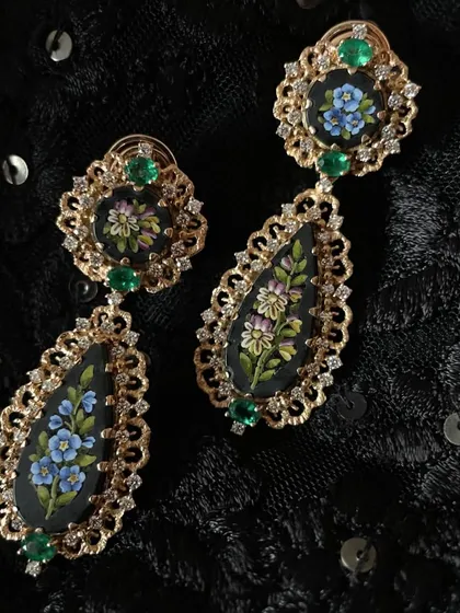 A pair of asymmetrical floral earrings with a vintage feel. The micromosaic flowers are set against a black background, framed by an ornate gold design with emeralds and diamonds.