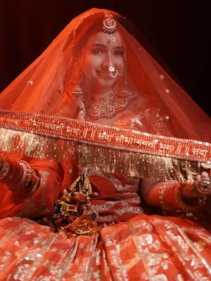 A beautiful shot of a bride in an orange-red lehenga, holding up her dupatta which has the 'Sada Saubhagyavati Bhava' blessing embroidered on it.